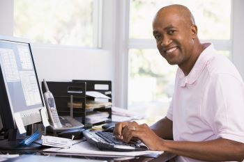 Man sitting at desk in home office, using keyboard and smiling with papers and mail sitting nearby. 