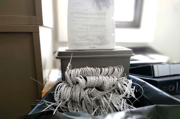 A sheet of paper being fed into an office paper shredder that is already overloaded with shredded paper coming out and piling up.