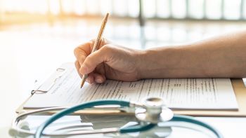 Hand holding a pen and writing on a document on a clipboard. A stethoscope is in the foreground.