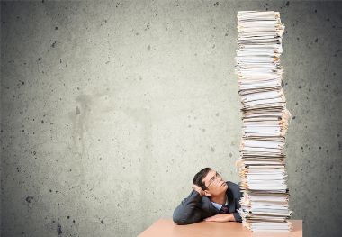 A man is sitting at a desk and looking up at a giant stack of papers and folders.