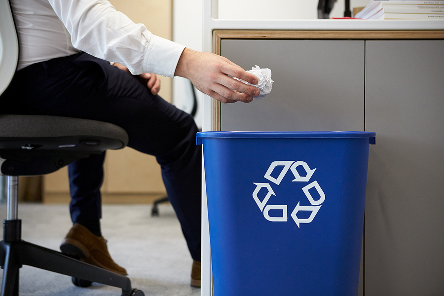 A person working in an office is dropping a crumpled sheet of paper into a recycling bin.