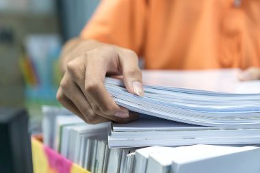 Office worker holding stack of documents waiting be managed on desk in busy office.